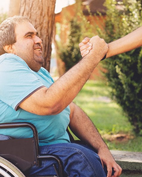 A man on a wheelchair, grabbing another person's hand.