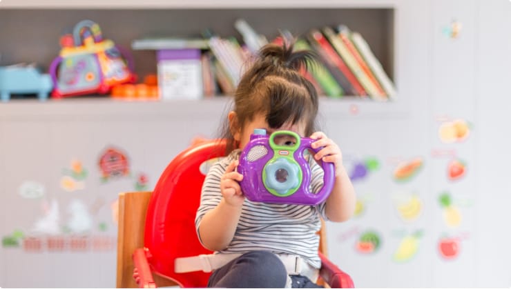 Young Girl Holding A Toy Camera To Her Face, Sitting In A Chair In A Classroom. Sensory and Behavioral Needs