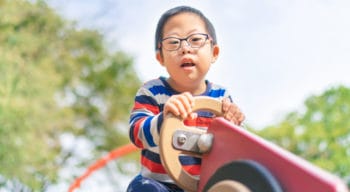 A small child with down syndrome is playing on a seesaw in a playground on a sunny day.
