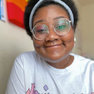 Devin, a young Black femme enby with a short brown fro, is shown chest-up smiling at the camera. They are wearing a white shirt, round glasses, and a gray headband.