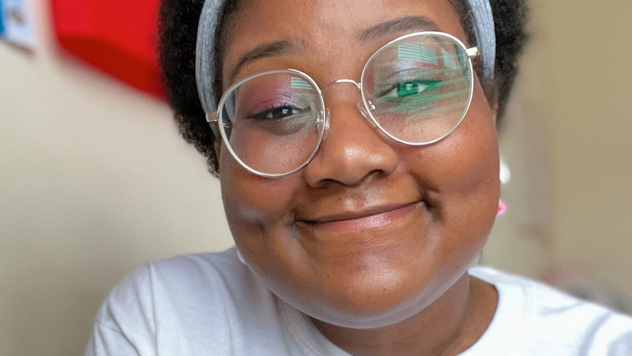 Devin, a young Black femme enby with a short brown fro, is shown chest-up smiling at the camera. They are wearing a white shirt, round glasses, and a gray headband.