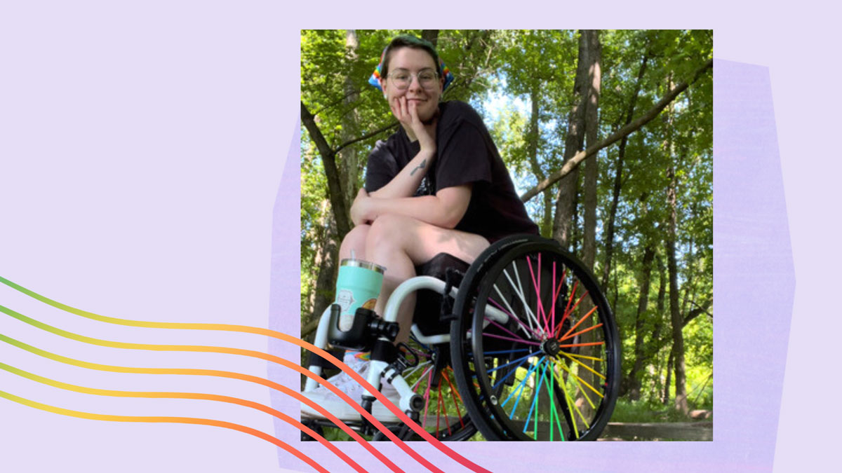 Em, a white femme presenting person, is sitting in front of a forest of trees in a wheelchair with a rainbow-spoked wheel.