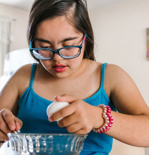 A teenage girl with Down syndrome cracking an egg into a clear bowl.