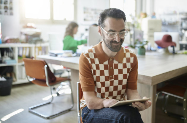 Close-up of Smiling Spanish design professional in early 40s relaxing in chair with digital tablet while colleagues work in background. support disability and neurodivergence