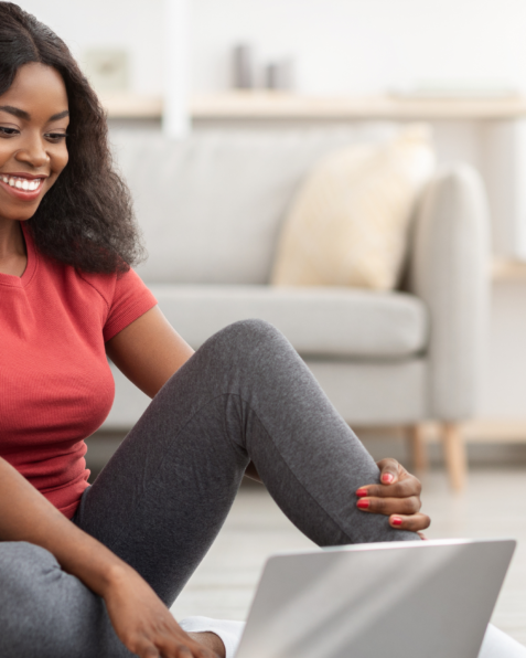 Women with black hair smiling in front of her laptop sitting on the floor.