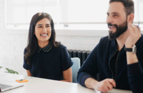 A smiling woman and a bearded man in a discussion at a bright office desk with a laptop.