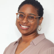 Portrait of Mesha Allen, a woman smiling with short black hair and orange glasses in a mauve blouse.