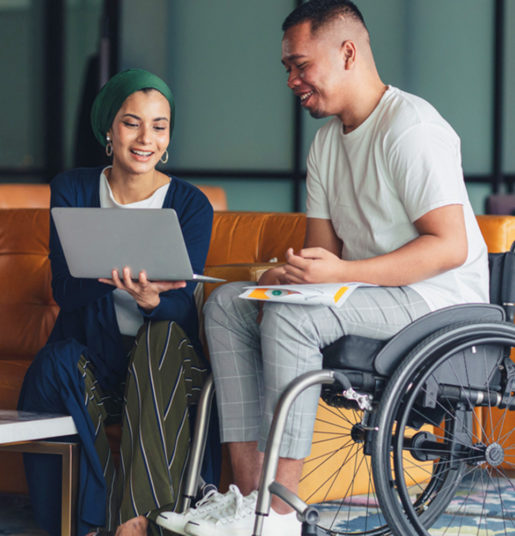 A man in a wheelchair looks at a laptop held by a woman on a couch