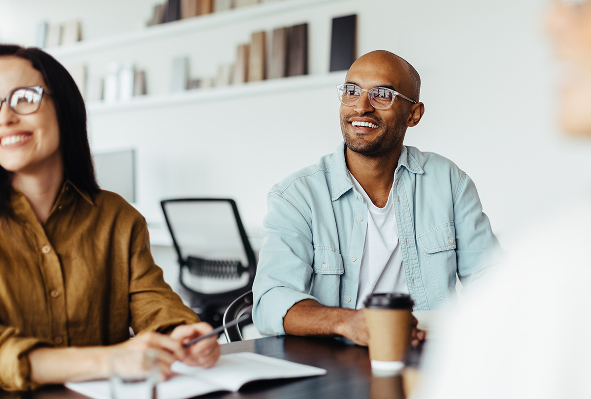 A man in glasses looks at someone talking during a meeting with coworkers.