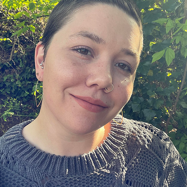 A smiling person with short hair, nose ring and earrings, and grey sweater.