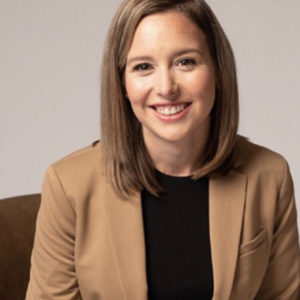 A woman with shoulder length hair smiles in a brown suit jacket and black shirt.