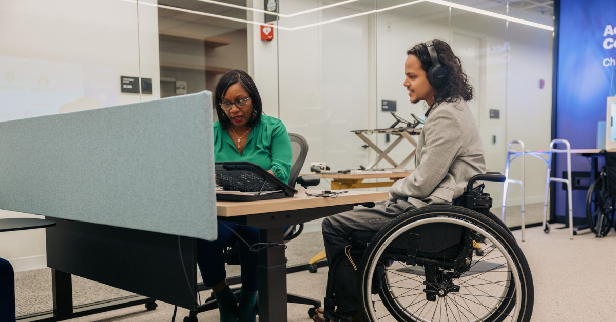 Two colleagues work together in an accessibility solutions center. One is wearing noise cancelling headphones and uses a wheelchair. The other reviews items with him on a laptop.