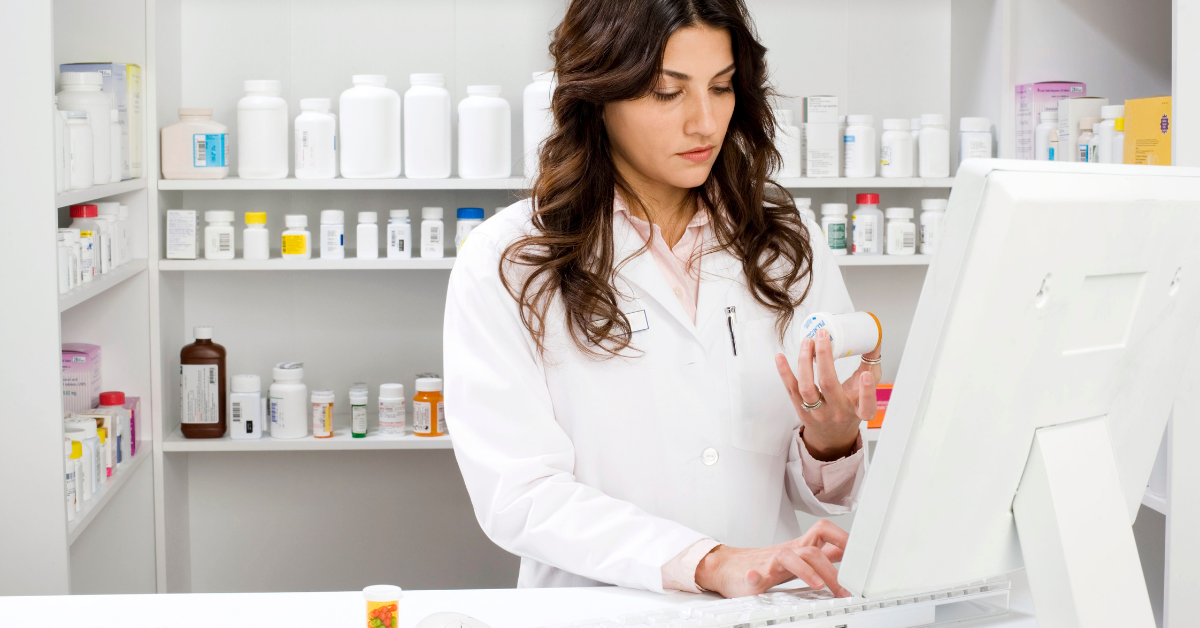 Long, dark haired pharmacist checking a prescription bottle at her computer. Behind her are shelves of different medications.