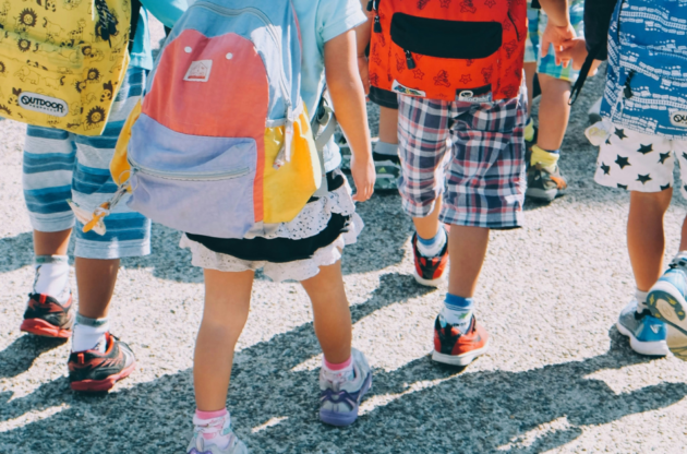 A group of children wearing colorful backpacks and casual clothing, walking on a sunlit pathway.