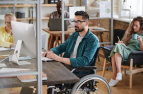 A man with glasses using a wheelchair is working in an office at his desktop iMAc computer. Coworkers out of focus are at their desks too.