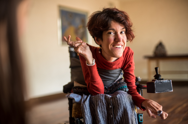 A woman with short reddish hair, a red long sleeve shirt and overalls sits in a wheelchair.