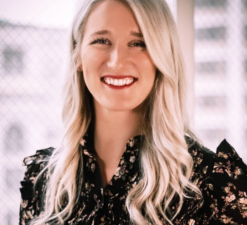 Portrait of Emily Hanson, a smiling woman with long blonde hair, wearing a patterned blouse, against a light background.