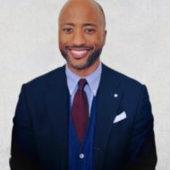 Portrait of Grant Harris, a smiling man in a dark suit with a red tie and a light background.