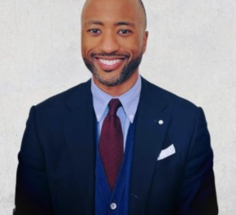 Portrait of Grant Harris, a smiling man in a dark suit with a red tie and a light background.