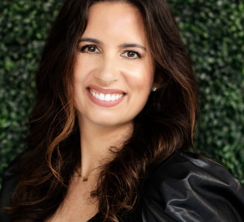Portrait of Melissa Danielsen, a smiling woman with long dark hair, wearing a black top, against a leafy green background.