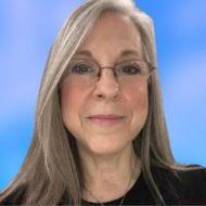 Portrait of Tracy Powell-Rudy, a smiling woman with glasses and long gray hair, wearing a black top, against a blue background.