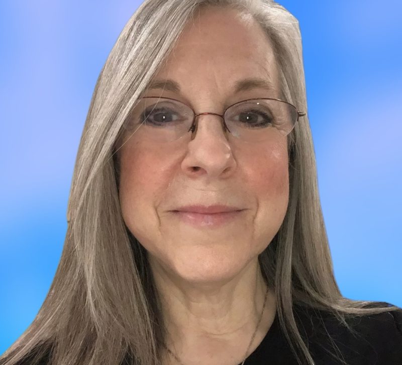 Portrait of Tracy Powell-Rudy, a smiling woman with glasses and long gray hair, wearing a black top, against a blue background.