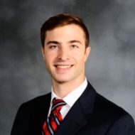 A man with short brown hair, wearing a dark suit and red striped tie, smiling against a gray background.