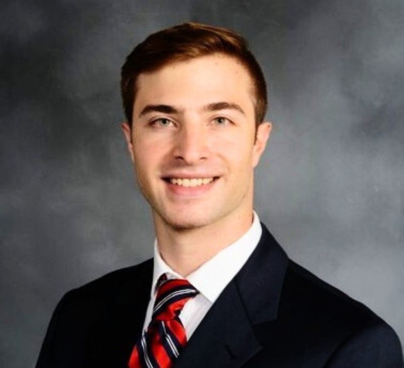 A man with short brown hair, wearing a dark suit and red striped tie, smiling against a gray background.