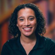 A woman with curly dark hair, wearing a black top, smiling against a colorful blurred background.