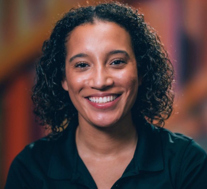 A woman with curly dark hair, wearing a black top, smiling against a colorful blurred background.