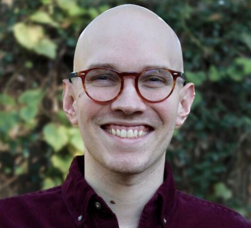 A man with round glasses, wearing a maroon shirt, smiling outdoors in front of greenery.