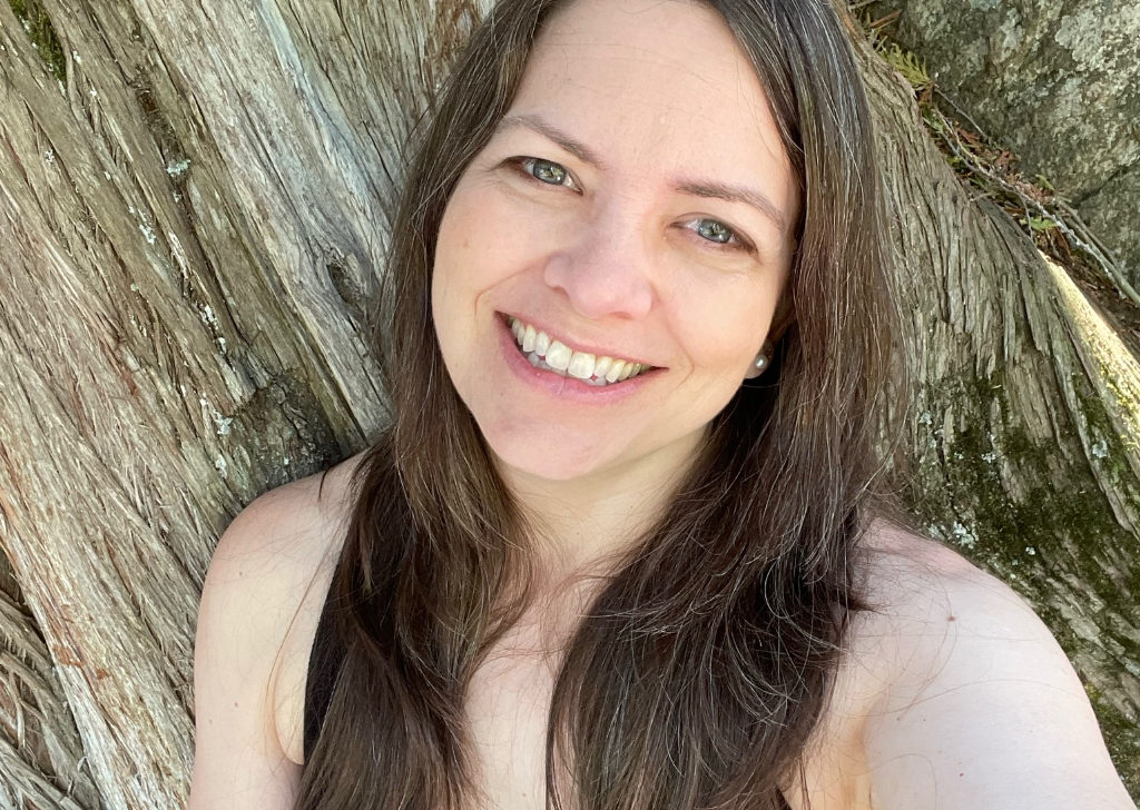 A woman smiling in front of a tree with brown hair in front of her shoulders.