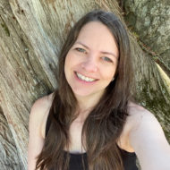 A woman smiling in front of a tree with brown hair in front of her shoulders.