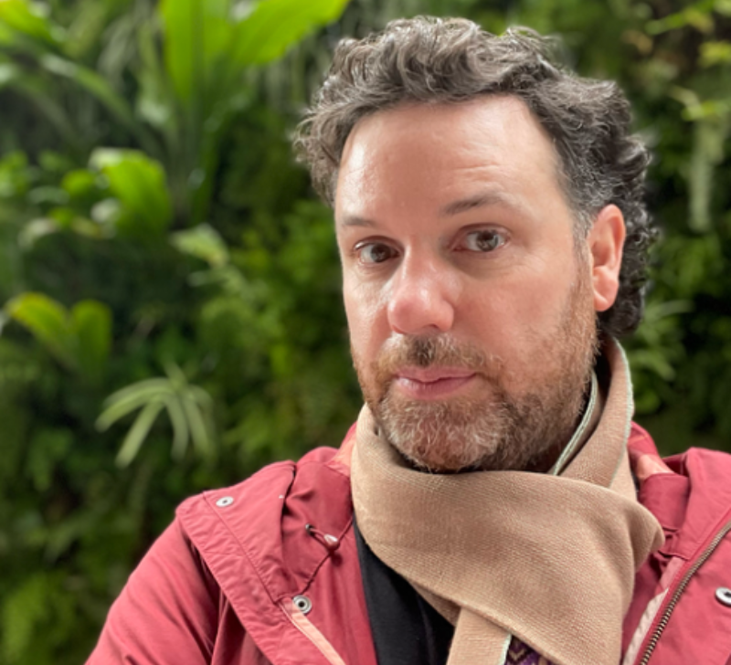 A man wearing a scarf and red jacket, seated in front of greenery, looking into the camera.