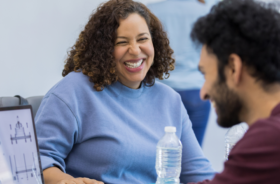 A woman with a blue sweater sits and laughs with a male colleague wearing a maroon sweater.