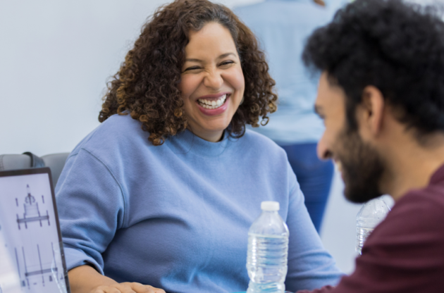 A woman with a blue sweater sits and laughs with a male colleague wearing a maroon sweater.
