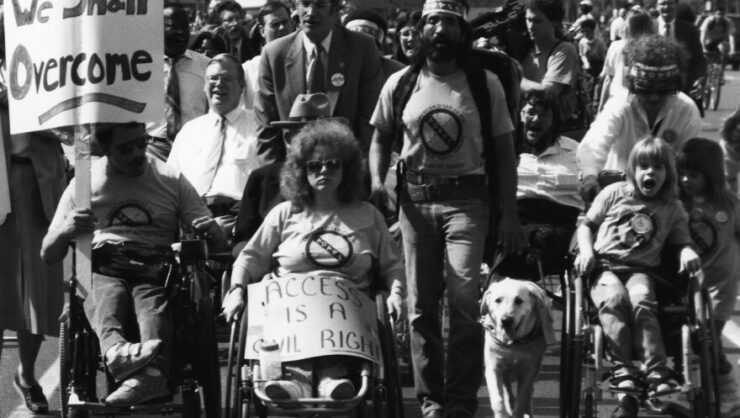 A black and white photo of a crowded group of disabled people marching for disability rights.